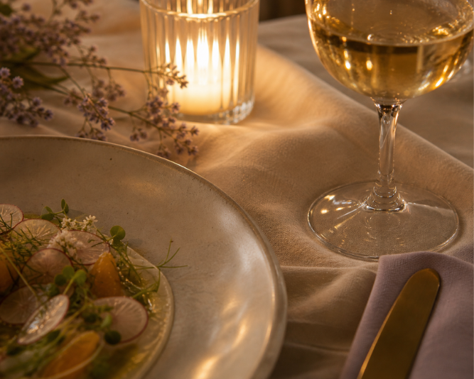A glass and the edge of a plate on ivory linen in candlelight, close and softly lit